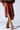 A person wearing a brown asymmetrical leather skirt and fur-trimmed ankle boots stands against a light background. The skirt, paired with the AZALEA WANG SHAINY MULTI MATERIAL SANDAL, features a front slit, while the boots are laced up with thick fur detailing, adding a touch of bold elegance to their ensemble.