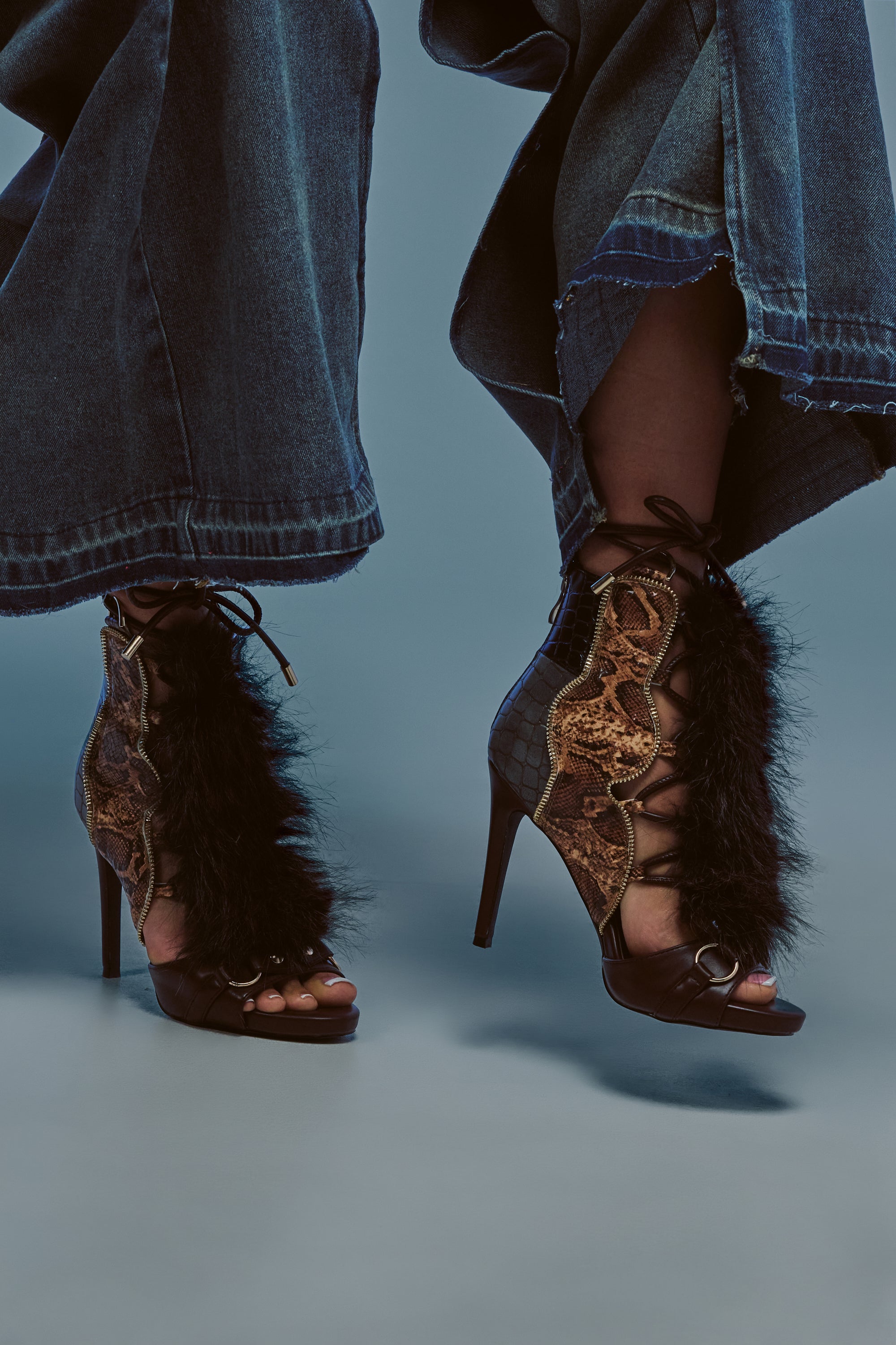 A person wearing wide-leg denim pants and the AZALEA WANG SHAINY SNAKE BROWN MULTI high heel shoes, featuring black fur and brown snakeskin patterns, standing on a light gray surface.