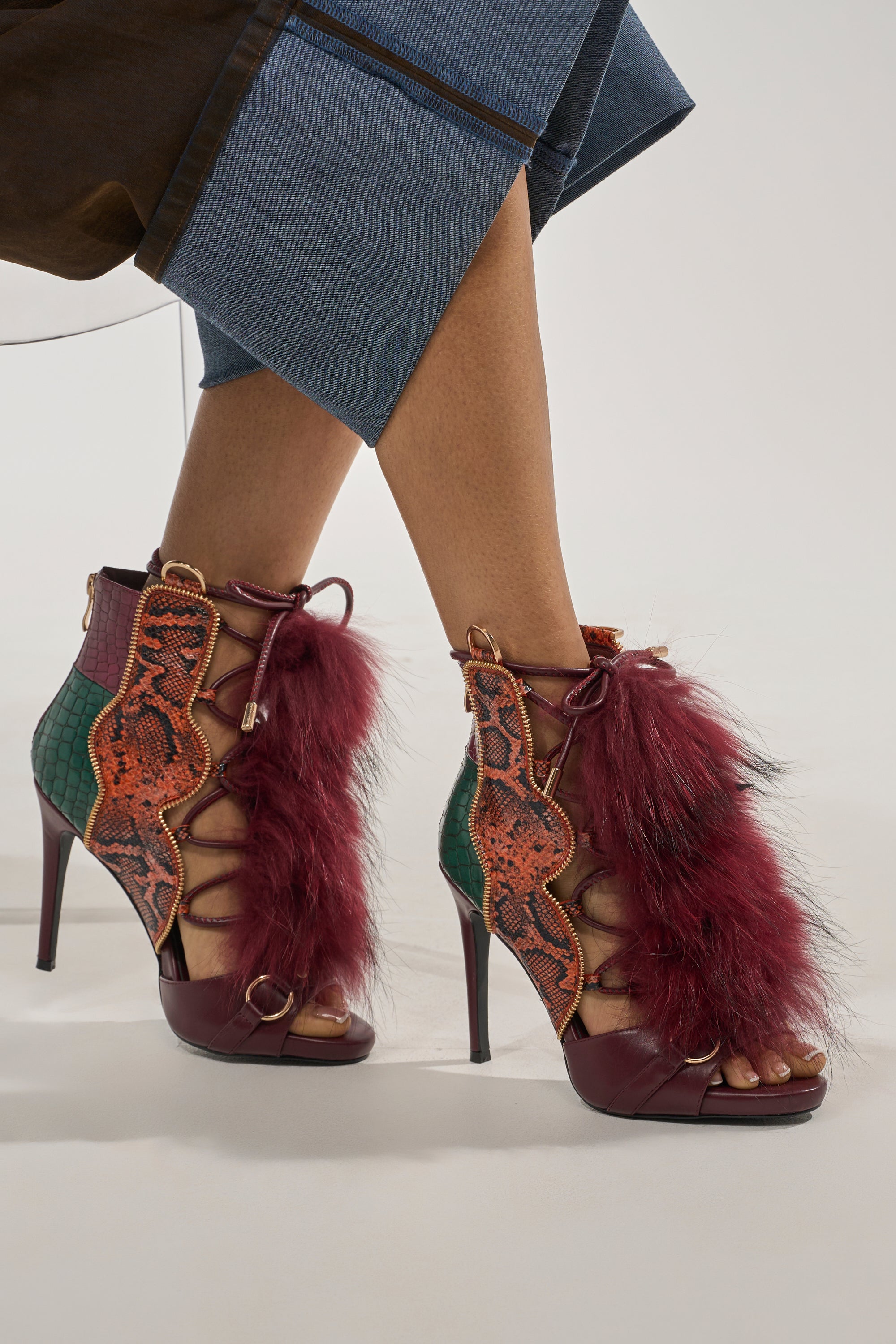 A person wearing the AZALEA WANG SHAINY SNAKE BURGUNDY MULTI high-heeled, open-toe lace-up boots with multicolored faux leather snakeskin panels, stiletto heels, large burgundy fur accents, paired with a denim skirt. 0