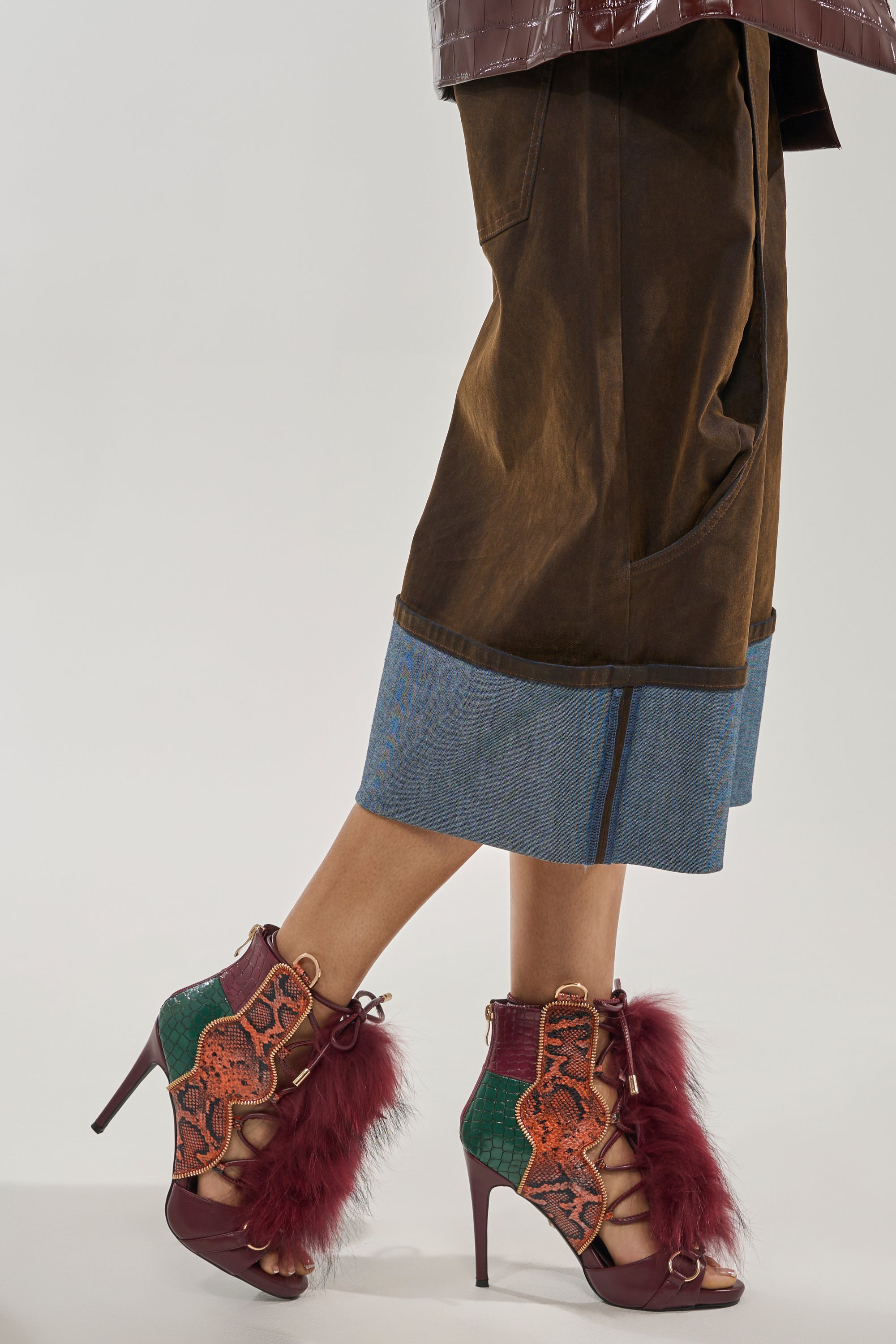 A person wears wide-legged brown pants with a denim cuff and AZALEA WANG SHAINY SNAKE BURGUNDY MULTI sandals, featuring burgundy heels, green and orange details, and fluffy maroon accents. 1