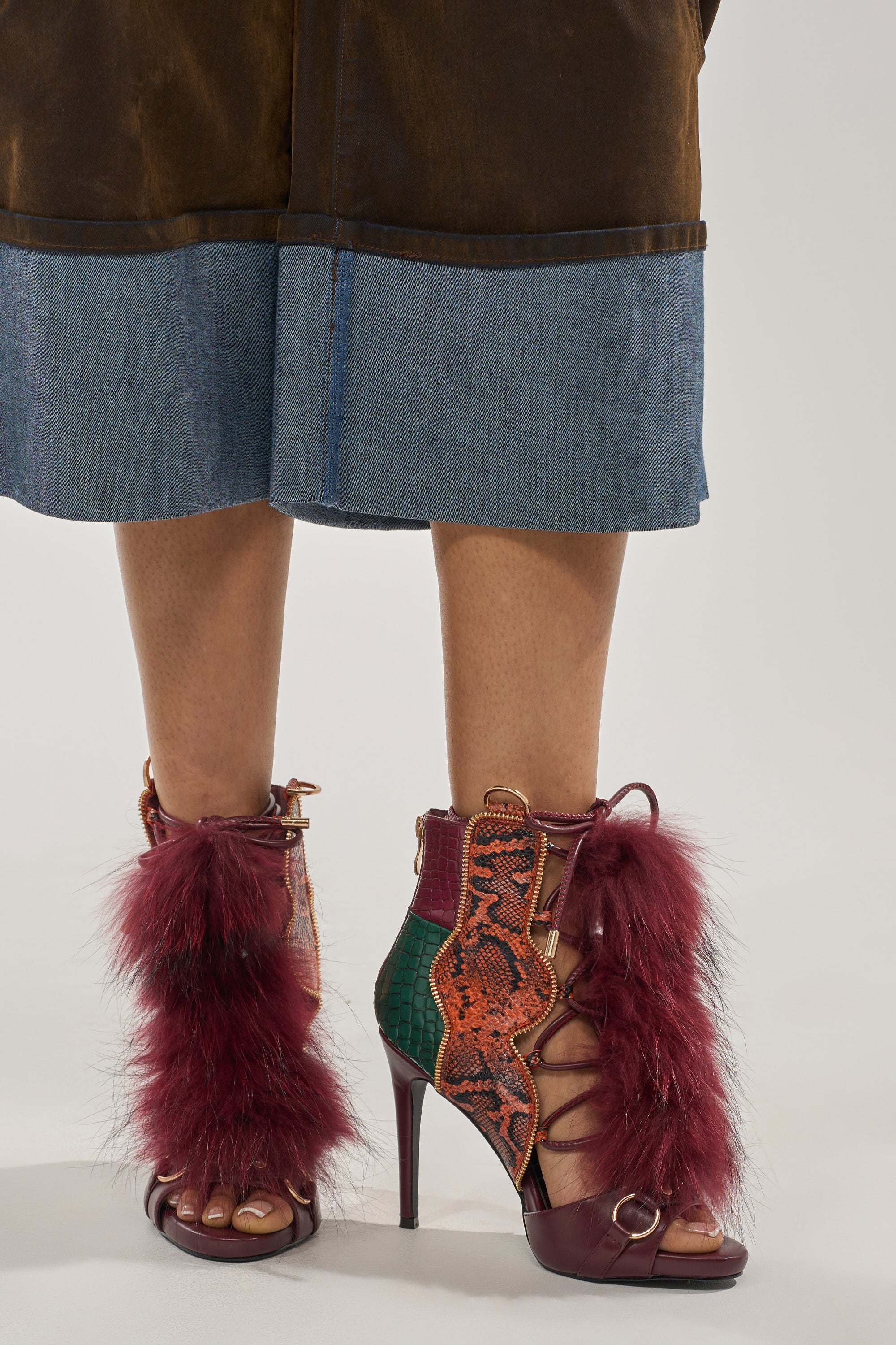A person in a brown and blue skirt wears AZALEA WANG SHAINY SNAKE BURGUNDY MULTI open-toe stiletto sandals with red faux fur, gold hardware, green snake print leather, and a lace-up front. 6