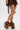 A person stands with legs crossed against a plain white background, wearing the AZALEA WANG SHAINY TAN FUR SANDAL and a brown lace skirt.