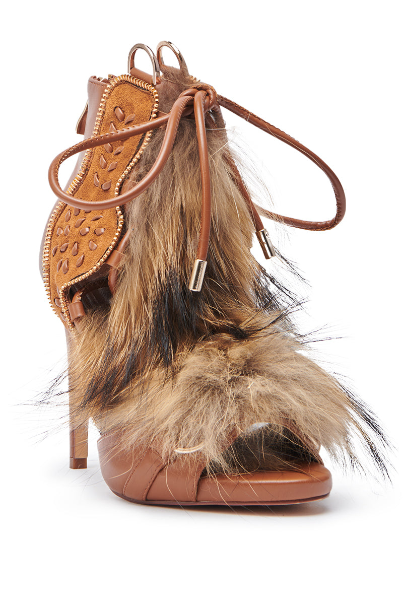 The AZALEA WANG SHAINY TAN FUR SANDAL is a brown high-heeled sandal with textured faux leather, gold zippers, laces, an ankle-high shaft, and bold tan and black faux fur patches on the upper and front strap.