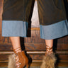 Indoors against a wooden background, someone wears wide-legged brown pants with light blue cuffs and the AZALEA WANG SHAINY TAN FUR SANDAL, a western-inspired tan lace-up shoe adorned with faux fur.