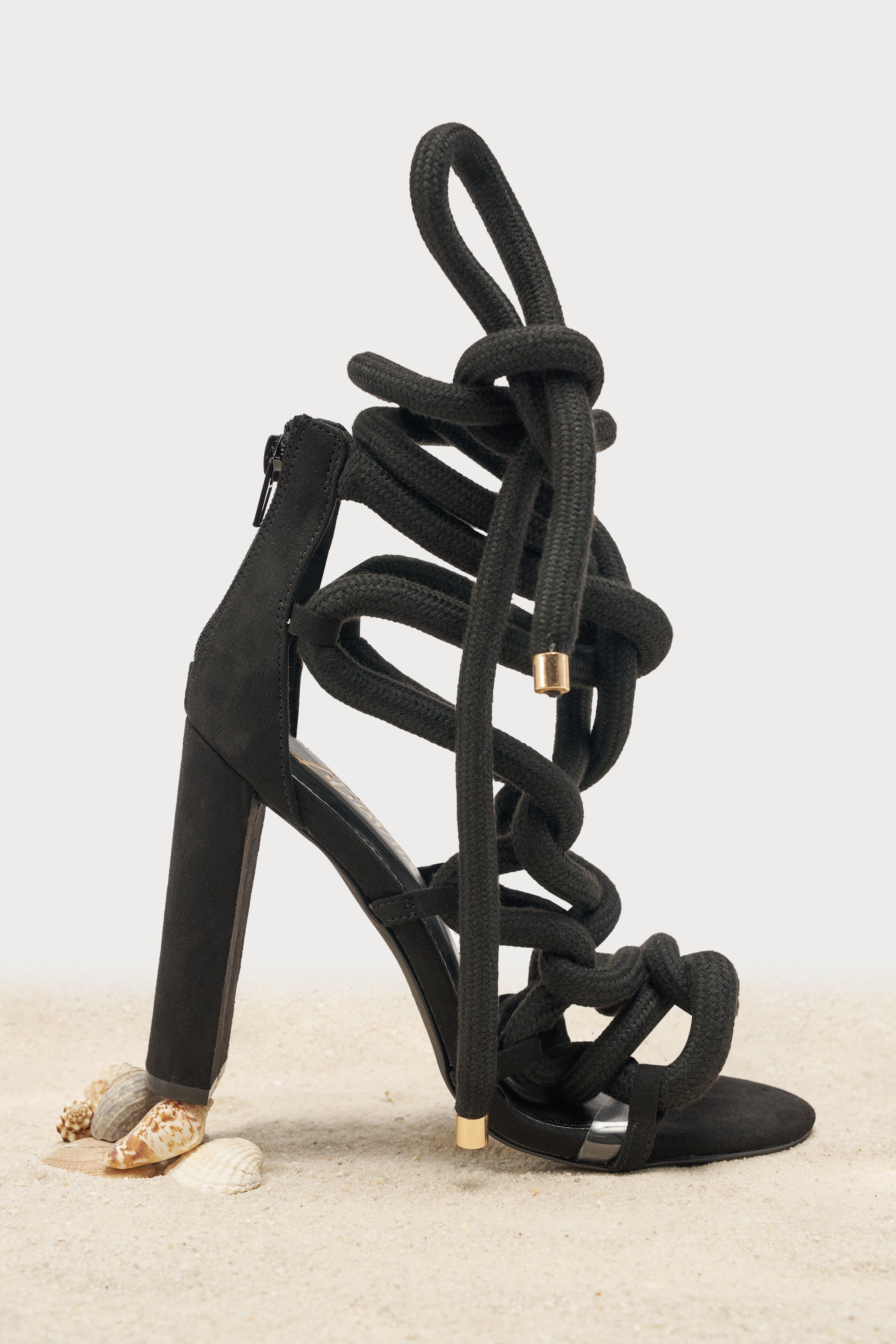 The AKIRA GOLD BLACK ROPE STRAP STILETTO SANDAL features black suede, thick crisscross rope straps twisted around the ankle and foot, styled with seashells on sand by the heel. 1