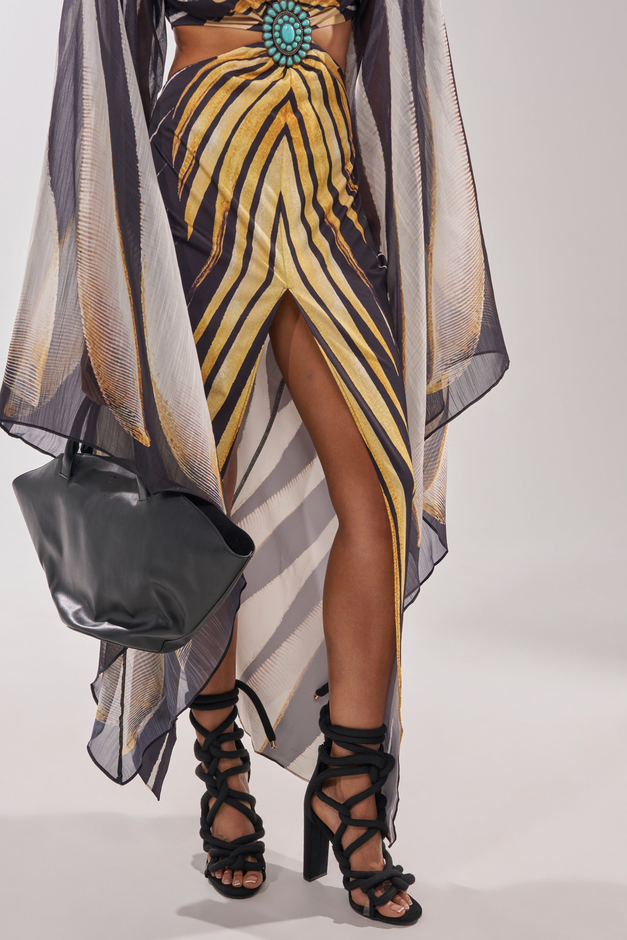 A person wearing a yellow and black geometric-patterned skirt with a high front slit, a sheer patterned cover-up, a turquoise belt, holding a black clutch bag, and AKIRA GOLD BLACK ROPE STRAP STILETTO SANDALS. 2