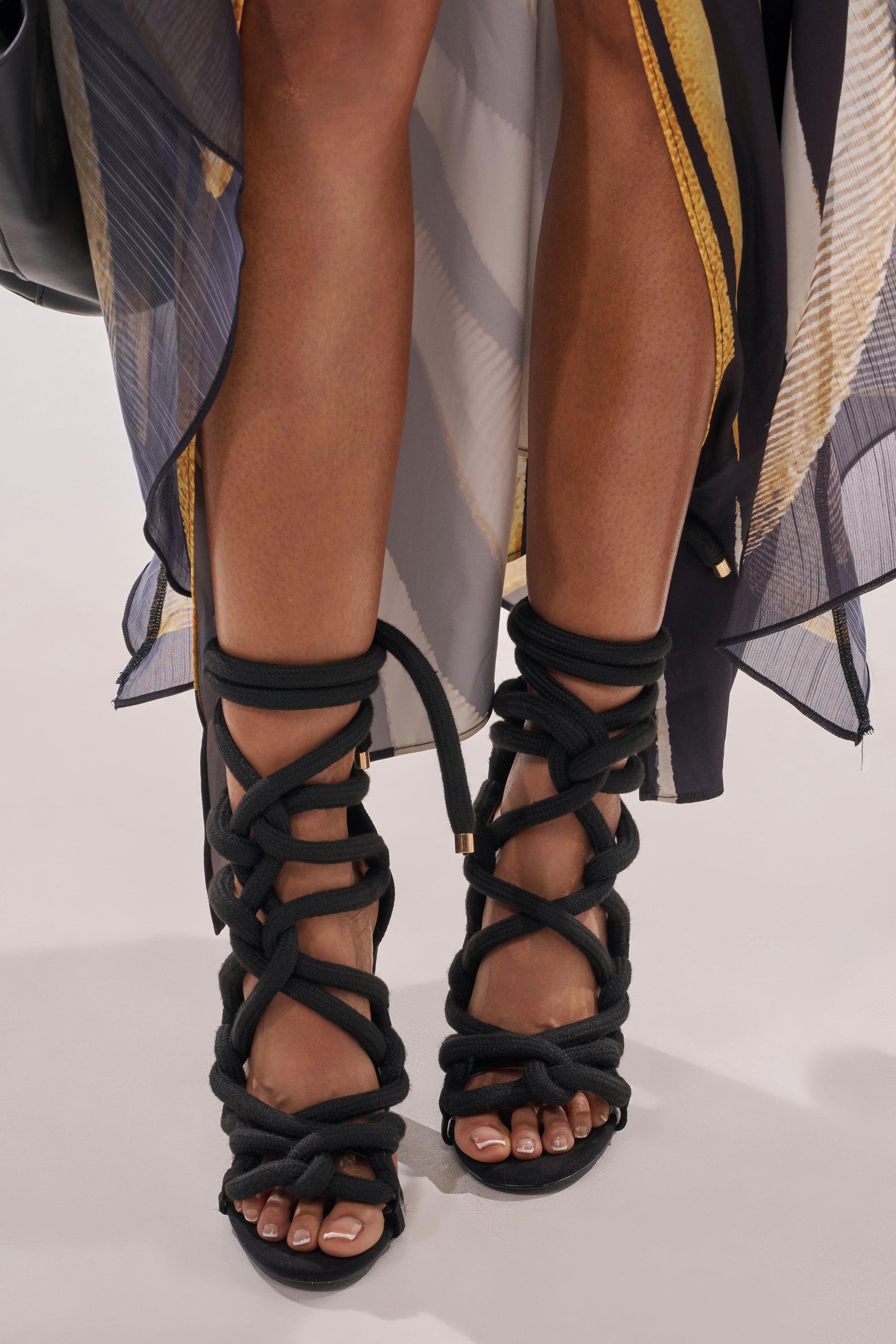 A close-up of a person wearing the AKIRA GOLD BLACK ROPE STRAP STILETO SANDAL features black suede stilettos with thick, braided rope straps. Paired with a patterned sheer skirt, their neutral-polished toes are visible through the design. 3