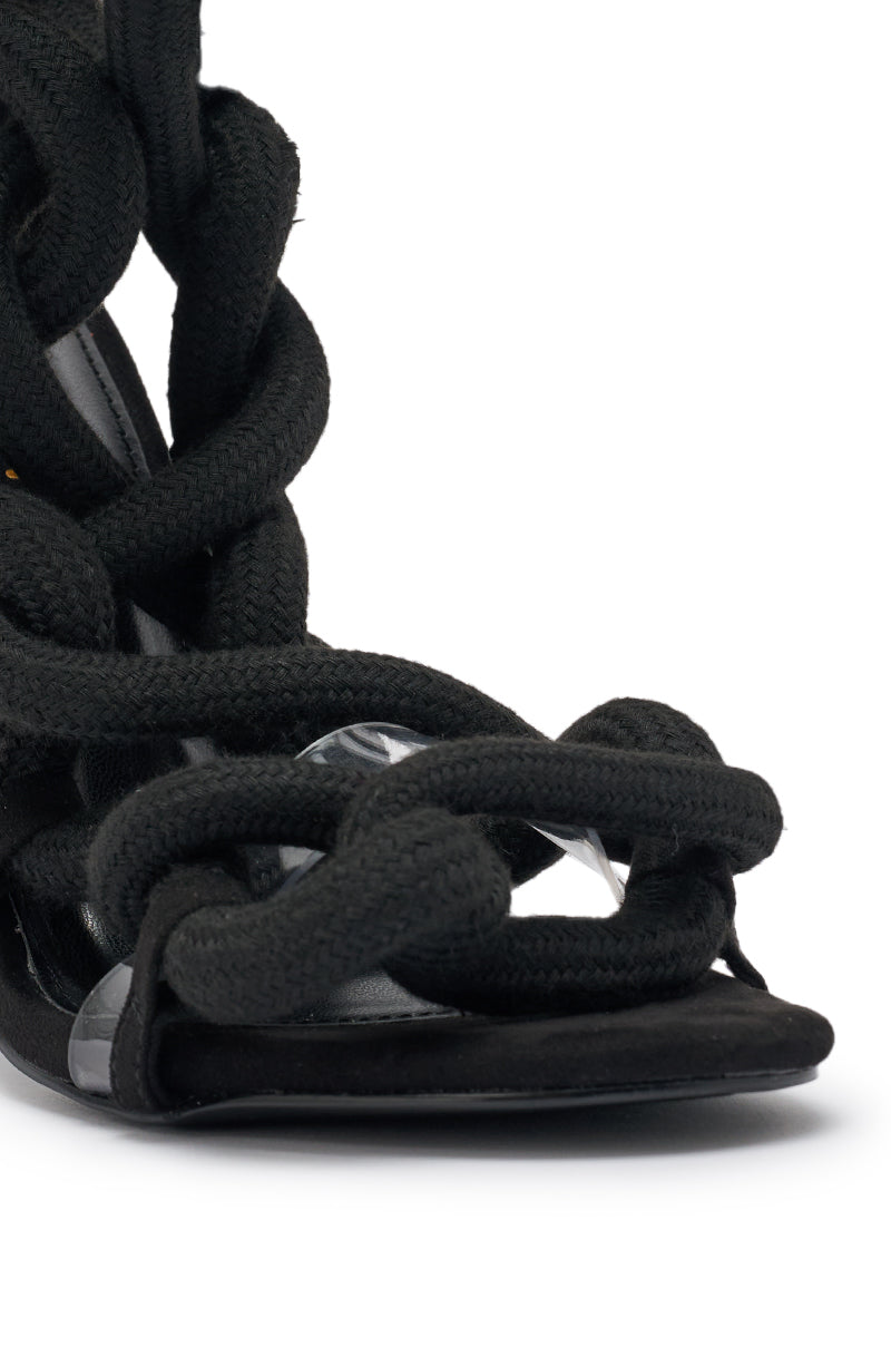 The AKIRA Gold Black Rope Strap Stiletto Sandal features a close-up of black suede, thick braided rope-like straps twisted across the front and sides, plus a clear accent near the sole. 7
