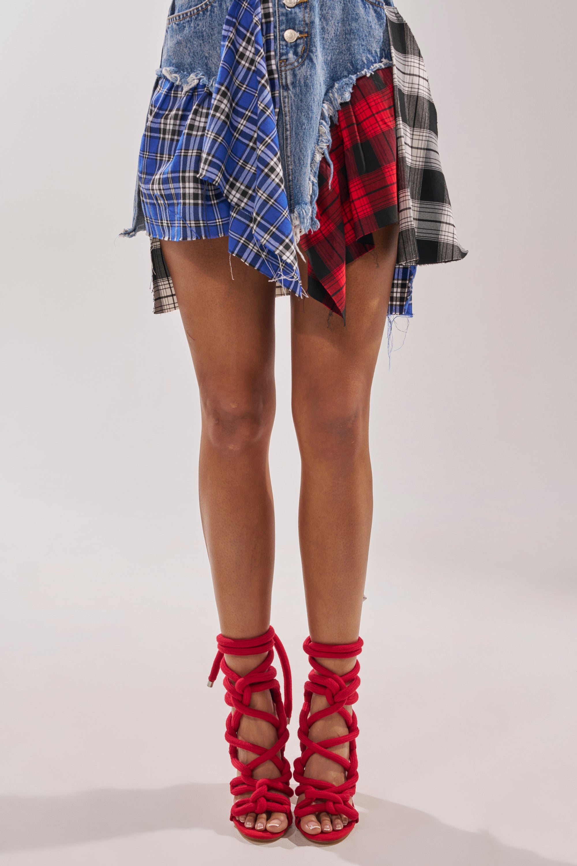 A person wearing a patchwork denim and plaid skirt pairs it with the AKIRA GOLD RED ROPE STRAP SANDAL, standing against a plain light background. 2