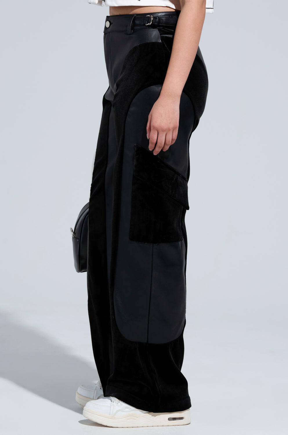 A person is wearing the OUTSIDE THE LINES PU PANT IN BLACK, which features high-waisted, wide-leg design with faux leather accents and cargo-style pockets. They complete this chic look with a white crop top and white sneakers, set against a neutral, light gray background.