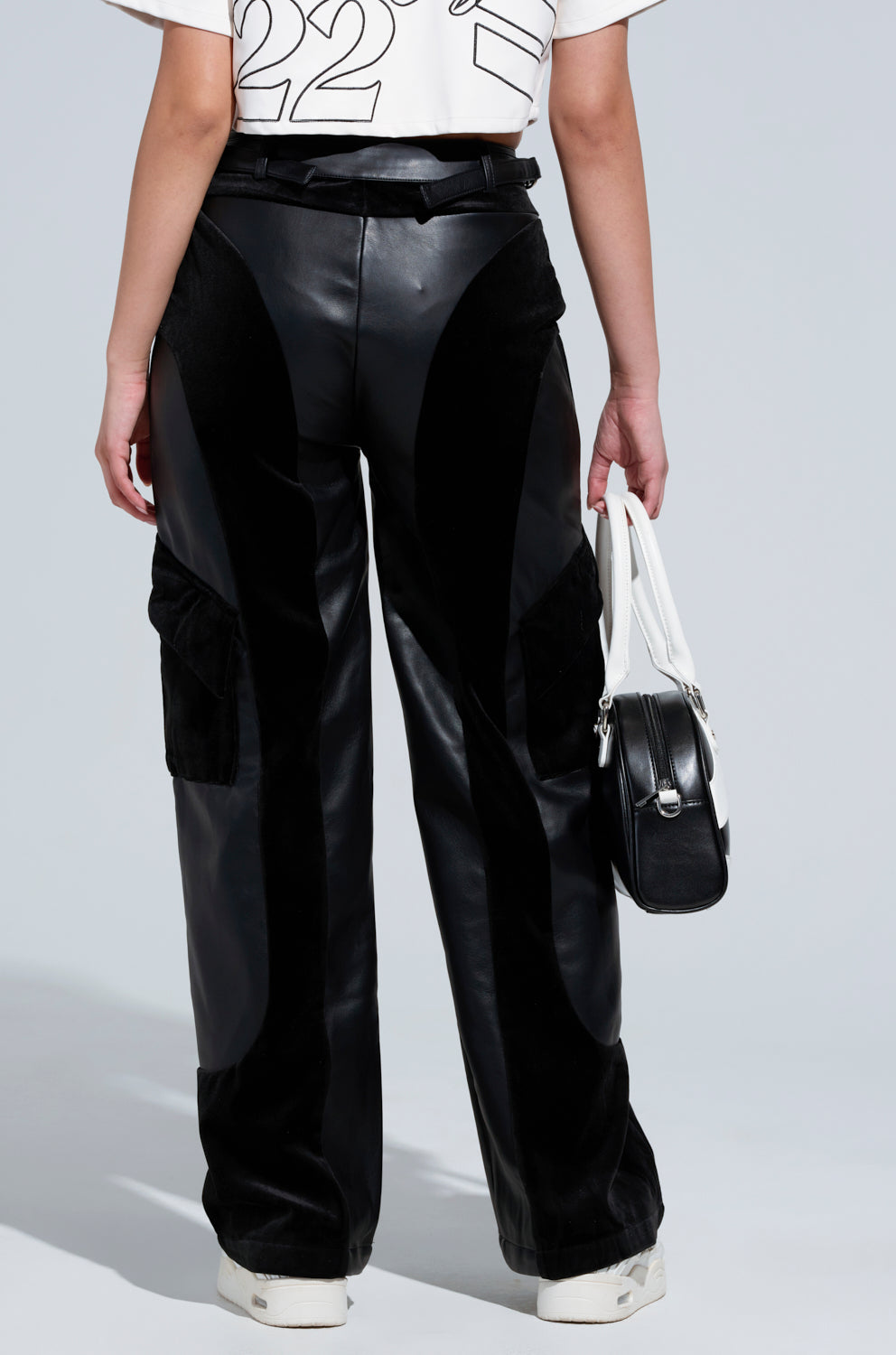 Wearing the sleek OUTSIDE THE LINES PU PANT IN BLACK with cargo-style pockets, a white graphic t-shirt, and matching white sneakers, the individual confidently showcases a black and white handbag against a neutral backdrop.
