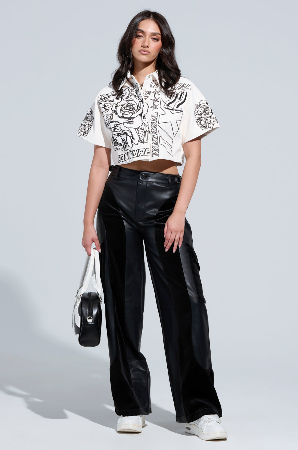 A person wearing a graphic crop top and the OUTSIDE THE LINES PU PANT IN BLACK holds a white handbag. They have long dark hair and are standing against a plain background, wearing white sneakers.