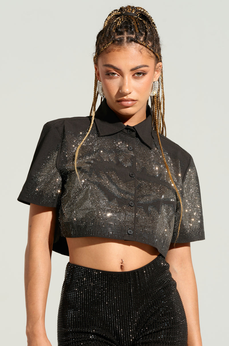 A person with long braided hair is wearing a shiny, black, short-sleeved MYSTERY LADY BUTTON DOWN TOP from AKIRA Label and matching high-waisted pants adorned with silver rhinestones. They have an intense expression and are posed against a light background. 0