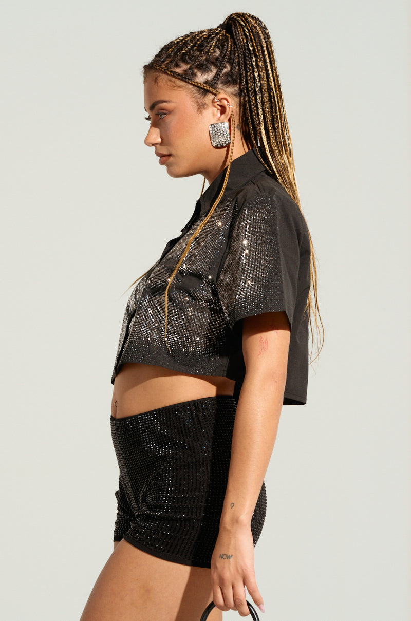 A person with long braided hair tied in a high ponytail stands in profile against a plain background. They wear the shiny black MYSTERY LADY BUTTON DOWN TOP adorned with silver rhinestones, paired with matching sequined shorts. They have a tattoo on their arm and are adorned with large, square, textured earrings. 2