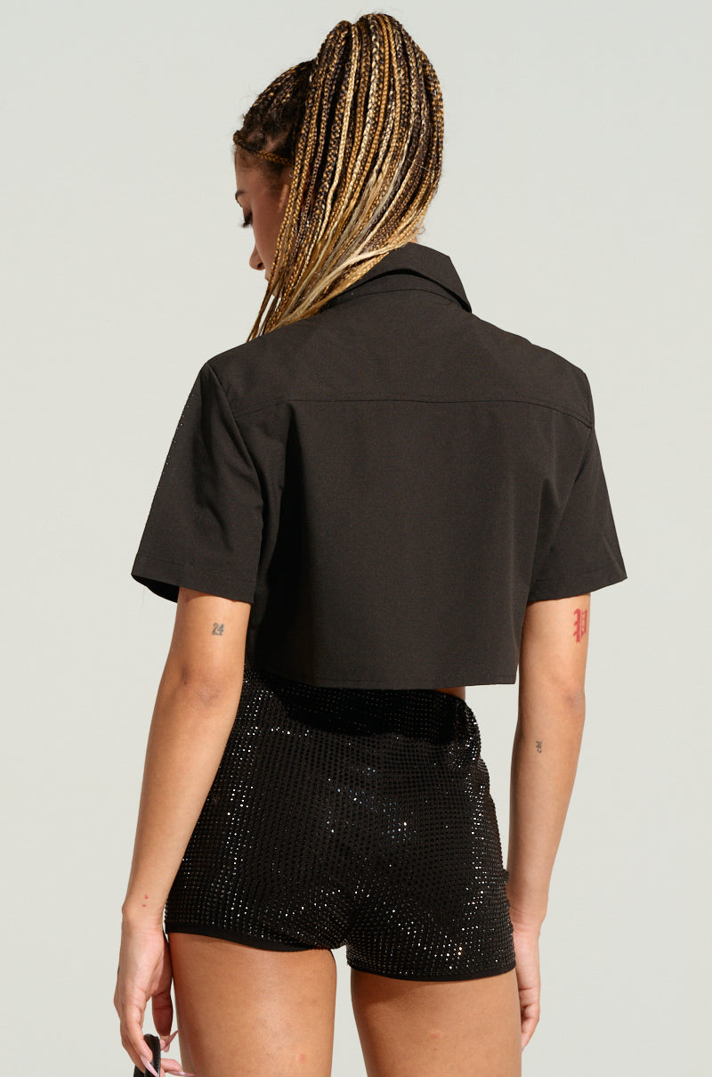A person with long braided hair is seen from behind, wearing the MYSTERY LADY BUTTON DOWN TOP, a short-sleeved, cropped black jacket adorned with silver rhinestones and shiny black shorts. The background is a plain, light-colored backdrop. 3