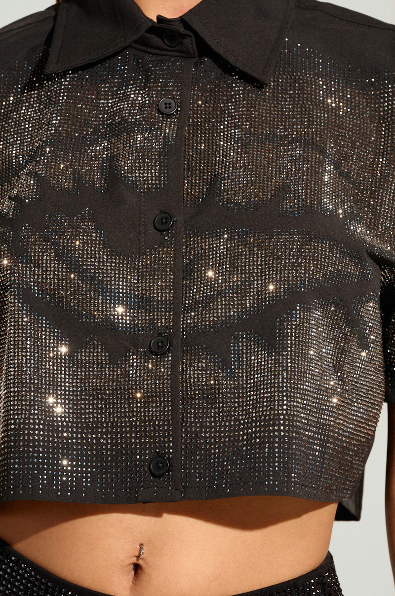 A close-up of a person wearing the MYSTERY LADY BUTTON DOWN TOP by AKIRA Label. This black, cropped top is adorned with sparkling silver rhinestones and features a collar and long sleeves. The bottom edge reveals the person's midriff and a small belly button ring, complemented by a subtle female eye design. 5