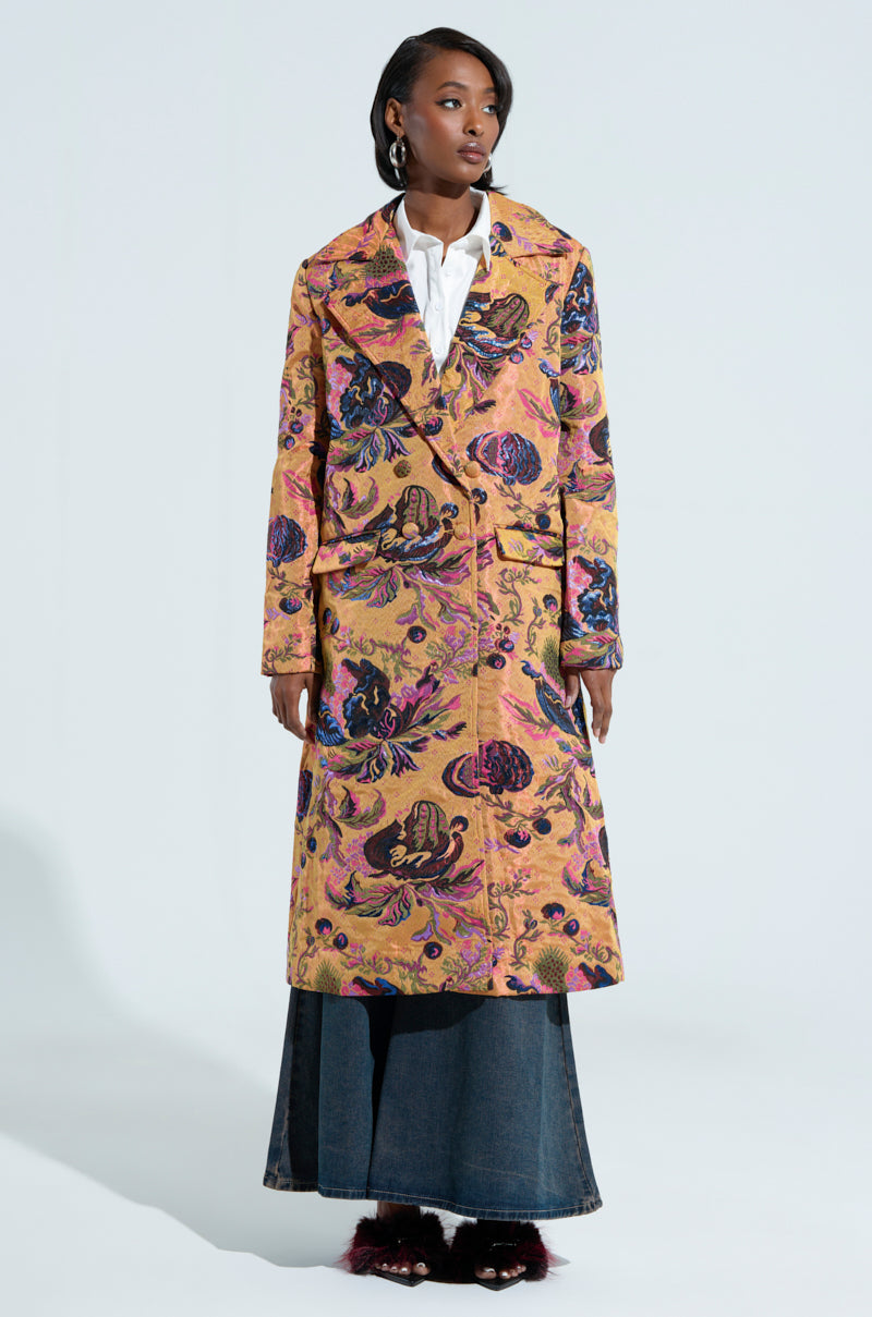 A person stands confidently against a plain background, clad in the CAMBRIDGE BROCADE TRENCH over a white shirt and dark denim skirt. The coat features an oversized fit and is alive with vibrant pink, purple, and gold hues. They sport short black hair and gaze resolutely to the side.