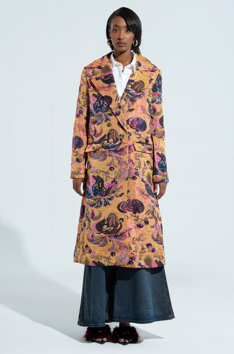 A person stands wearing the CAMBRIDGE BROCADE TRENCH over a white shirt and wide-leg denim pants. They have dark hair and pose against a plain backdrop, showcasing the vibrant pattern of the coat.
