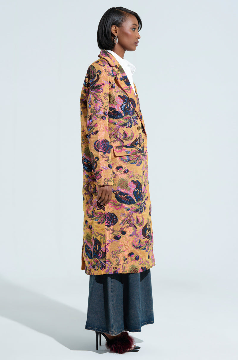 A person is shown from the side, dressed in a CAMBRIDGE BROCADE TRENCH featuring intricate purple and gold patterns. They pair it with wide-legged jeans and dark heels, while their shoulder-length hair and earrings add to the outfit's elegance against a simple backdrop.
