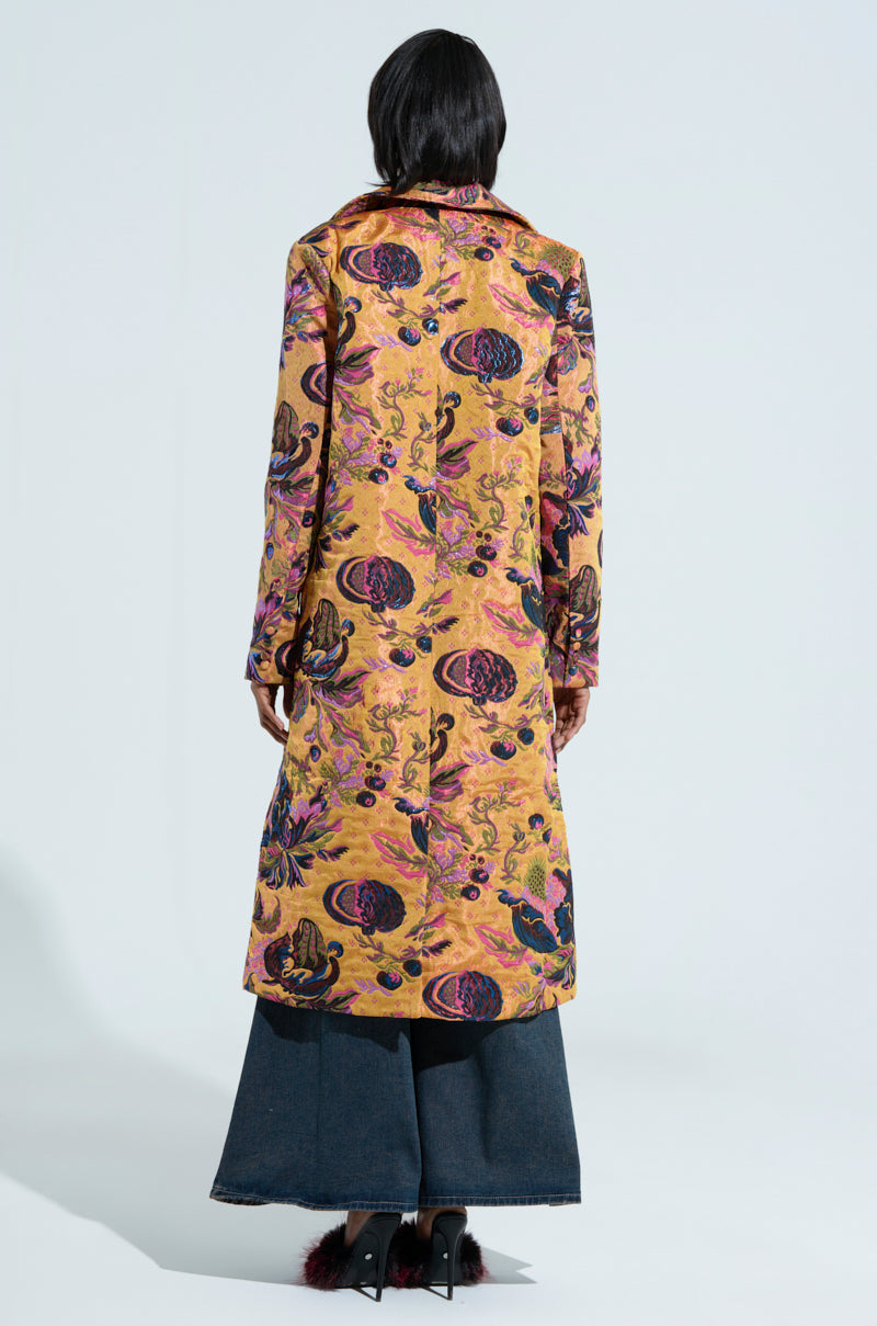 A person with long dark hair is dressed in the CAMBRIDGE BROCADE TRENCH, showcasing an elaborate floral design. The trench coat displays a vibrant combination of gold, purple, and red hues. They stand against a simple light background, with wide-legged jeans visible beneath the coat.