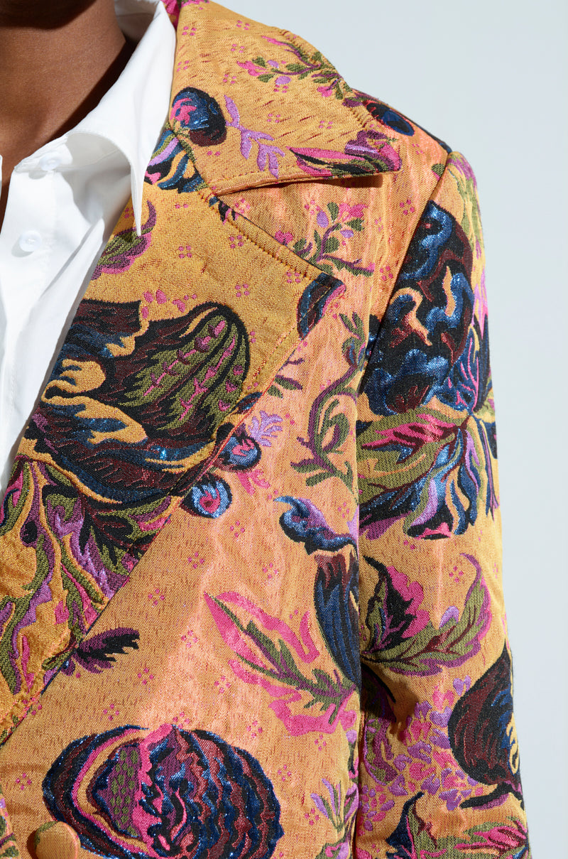 A close-up of someone wearing a blazer adorned with floral patterns in vivid colors such as pink, green, and blue over a white shirt. The intricate designs and textured fabric mirror the elegance of the CAMBRIDGE BROCADE TRENCH.