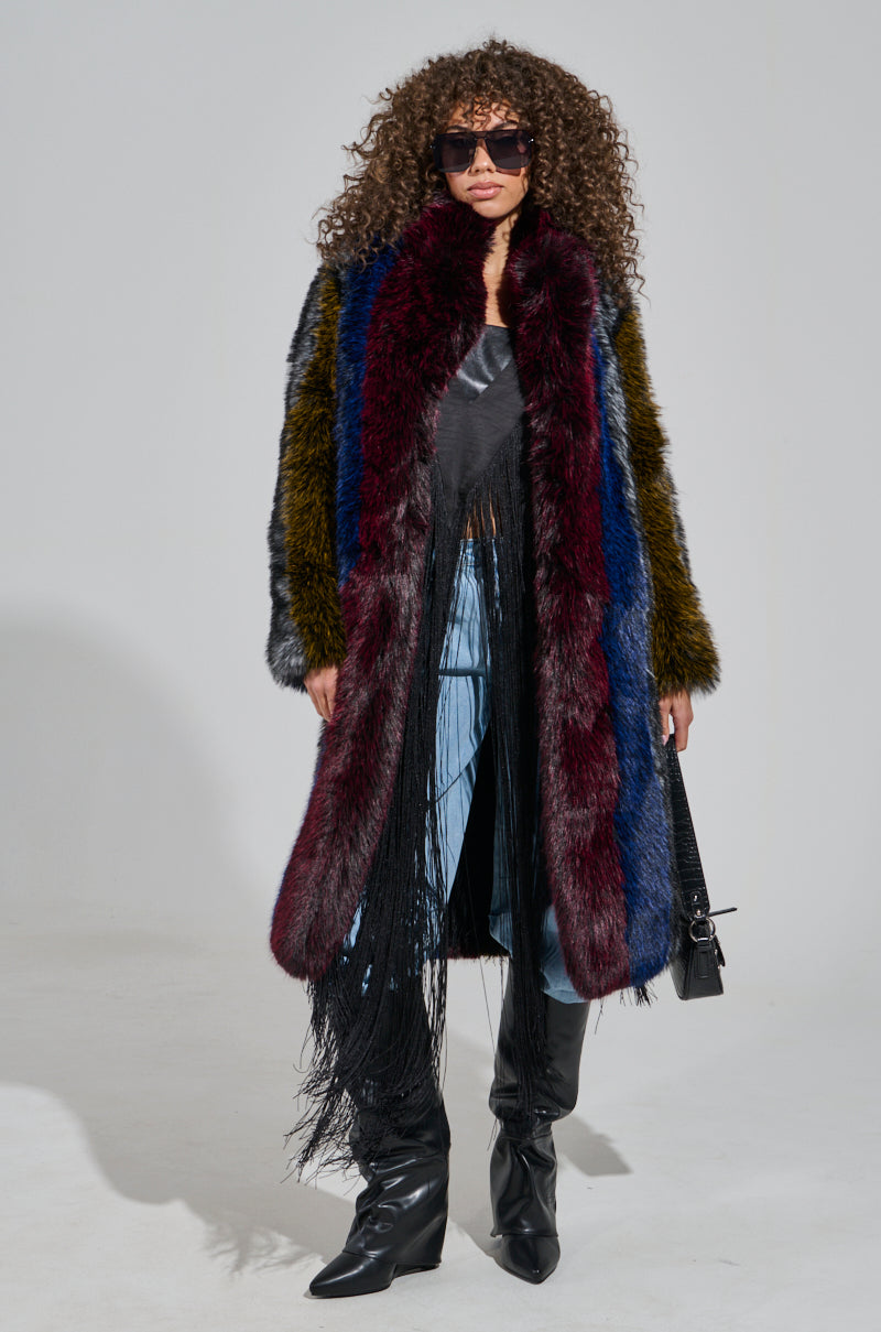A person with curly hair and sunglasses dons the luxurious SHIGO FAUX FUR TRENCH, featuring vibrant shades of burgundy, blue, and green. They complement it with a black top, fringed pants, knee-high boots, and carry a small handbag against a plain, neutral backdrop.