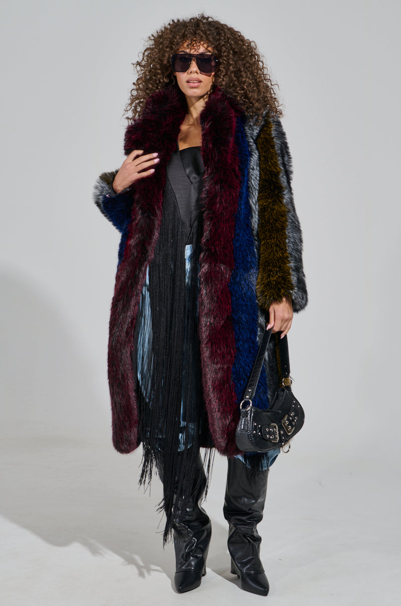 A person with curly hair and sunglasses exudes confidence in the SHIGO FAUX FUR TRENCH, a multi-colored faux fur statement piece. They pair it with a black fringed outfit, over-the-knee black boots, and hold a small black handbag. The plain white background accentuates their luxury fashion ensemble.