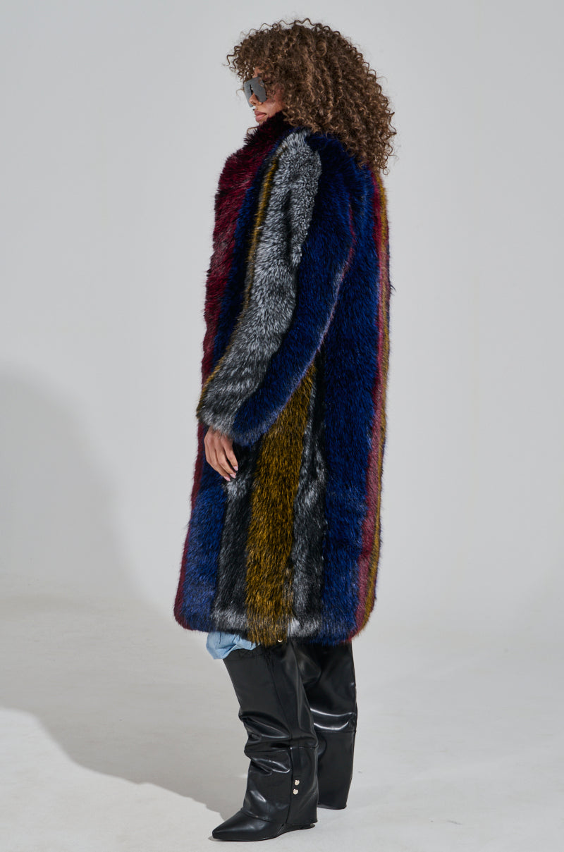 A person wearing the SHIGO FAUX FUR TRENCH, featuring a long and multicolored faux fur design with stripes of blue, red, and gray, stands in profile. Their curly hair complements the sunglasses and black knee-high boots they are accessorized with. This striking outerwear piece exemplifies luxury fashion against a plain white background.
