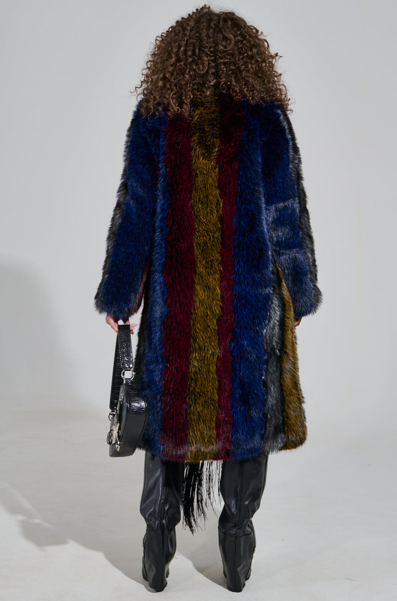 A person with curly hair is wearing the SHIGO FAUX FUR TRENCH, showcasing its luxurious design with vertical blue, red, and yellow stripes. They complement it with black knee-high boots and carry a black handbag, exuding confidence against a plain backdrop.