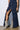 A person is wearing a dark denim skirt with multiple pockets and a front zipper, revealing part of their leg. They are also sporting the AZALEA WANG SIBYLLE BEIGE SNAKE COCKTAIL BOOTIE, which features high heels and a pointed toe with a faux snakeskin pattern. The background is a plain white wall.