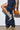 A person wearing a dark denim skirt with a high slit confidently showcases the AZALEA WANG SIBYLLE BEIGE SNAKE COCKTAIL BOOTIE. They hold a small, shiny gold handbag against the plain white wall backdrop.