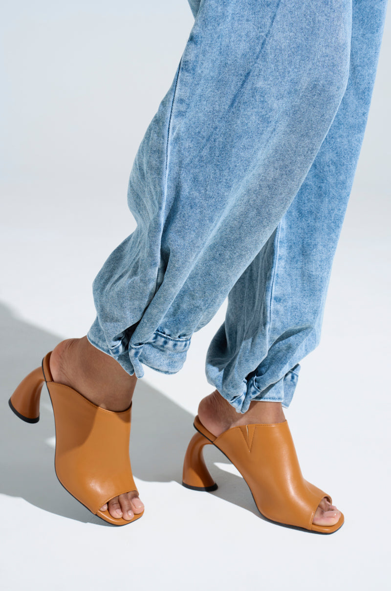 A person in light blue, loose-fitting jeans stands on a light surface wearing stylish AZALEA WANG tan slip-on mule sandals with chunky heels. The jeans are slightly rolled up at the ankles, showcasing the fashionable footwear.