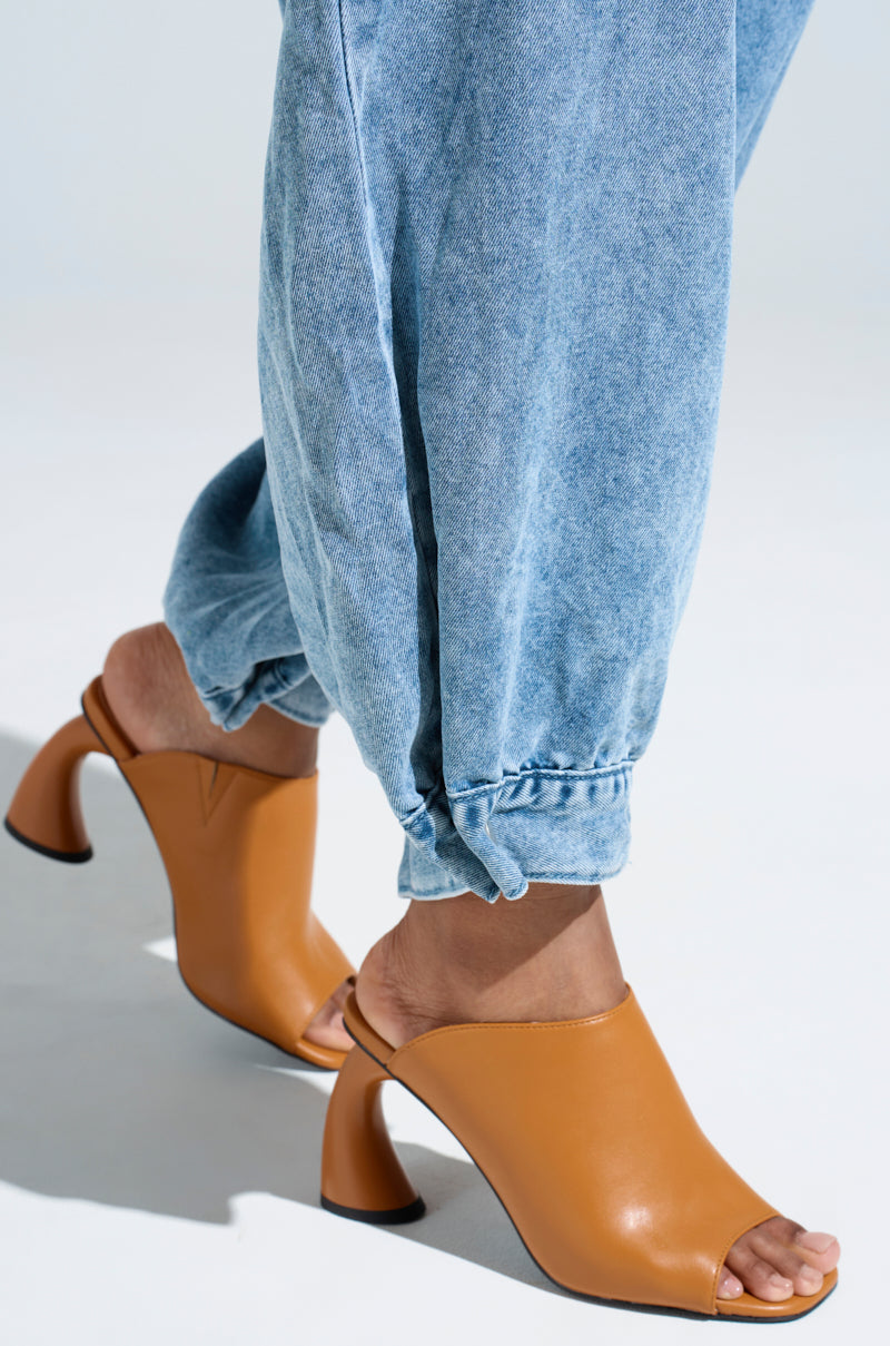 Wearing light blue denim pants and brown AZALEA WANG TAN SLIP ON MULE SANDAL, featuring a unique curved chunky heel. The slip-on vegan leather sandals add an eco-friendly touch to the chic look, all set against a light background.