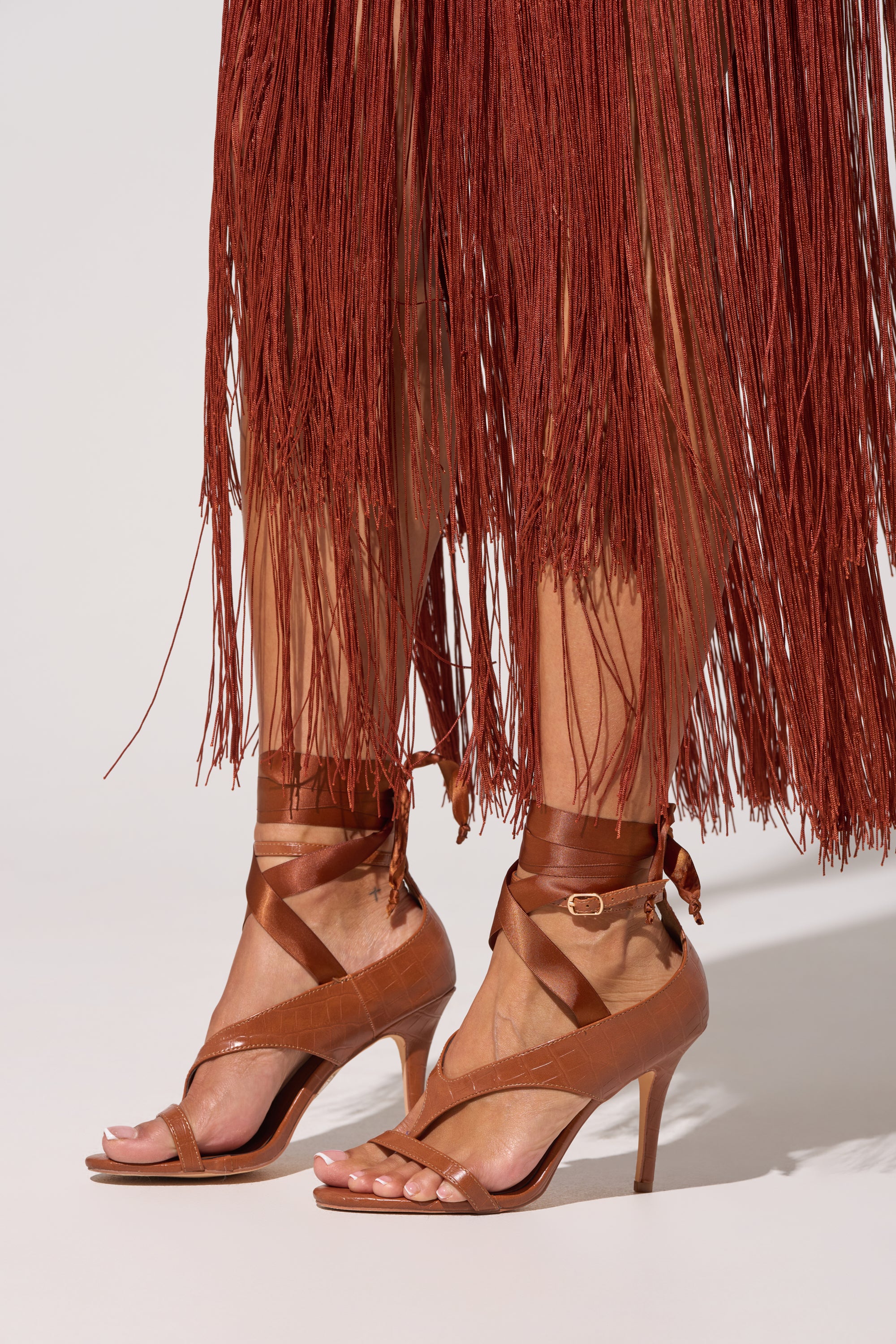 Close-up of a person wearing the AKIRA GOLD SIEGER BROWNPATENT WRAP UP BALLERINA STILETTO SANDAL with ankle ties and a comfort cushion insole, paired with a long rust-colored fringed skirt against a plain light background. 0