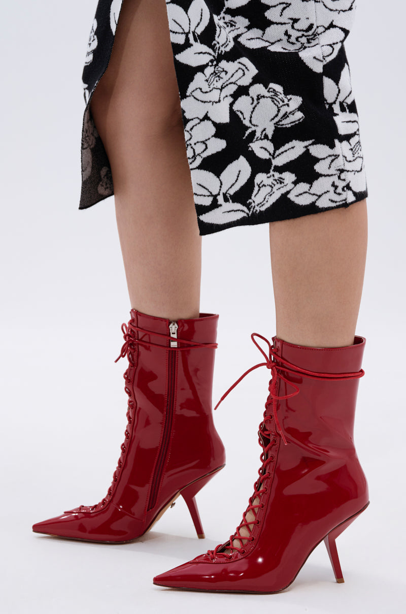 A person wearing a black and white floral skirt stands in shiny AZALEA WANG SILVA RED LACE UP STILETTO BOOTIES featuring pointed toes, thin sculptural stiletto heels, and a glossy faux patent leather finish.