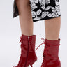 A person wearing a black and white floral skirt stands in shiny AZALEA WANG SILVA RED LACE UP STILETTO BOOTIES featuring pointed toes, thin sculptural stiletto heels, and a glossy faux patent leather finish.