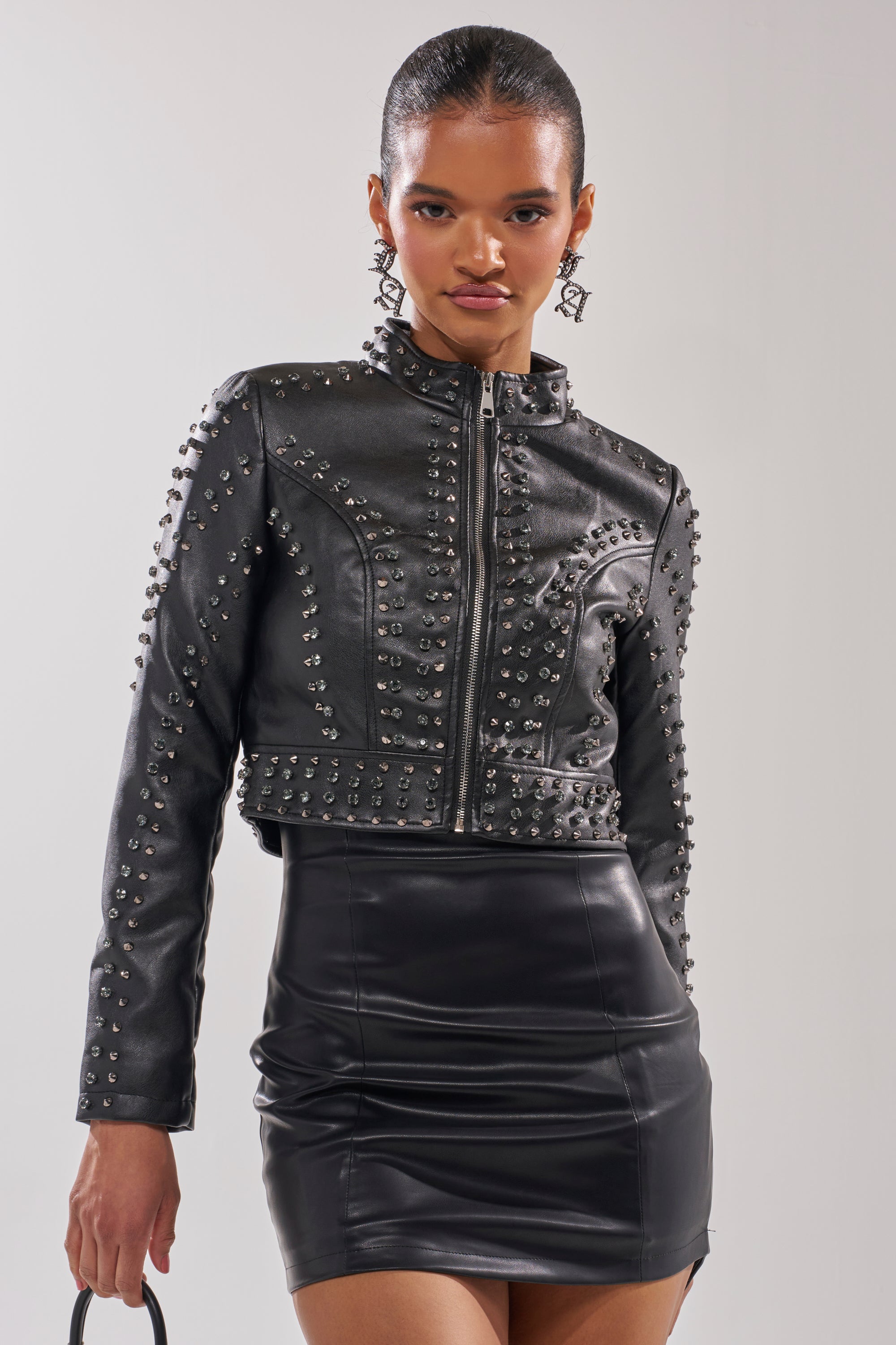 A woman confidently stands against a light background wearing the SYLVIE STUDDED MOTO jacket, a black leather mini skirt, and holding a small black bag. 0