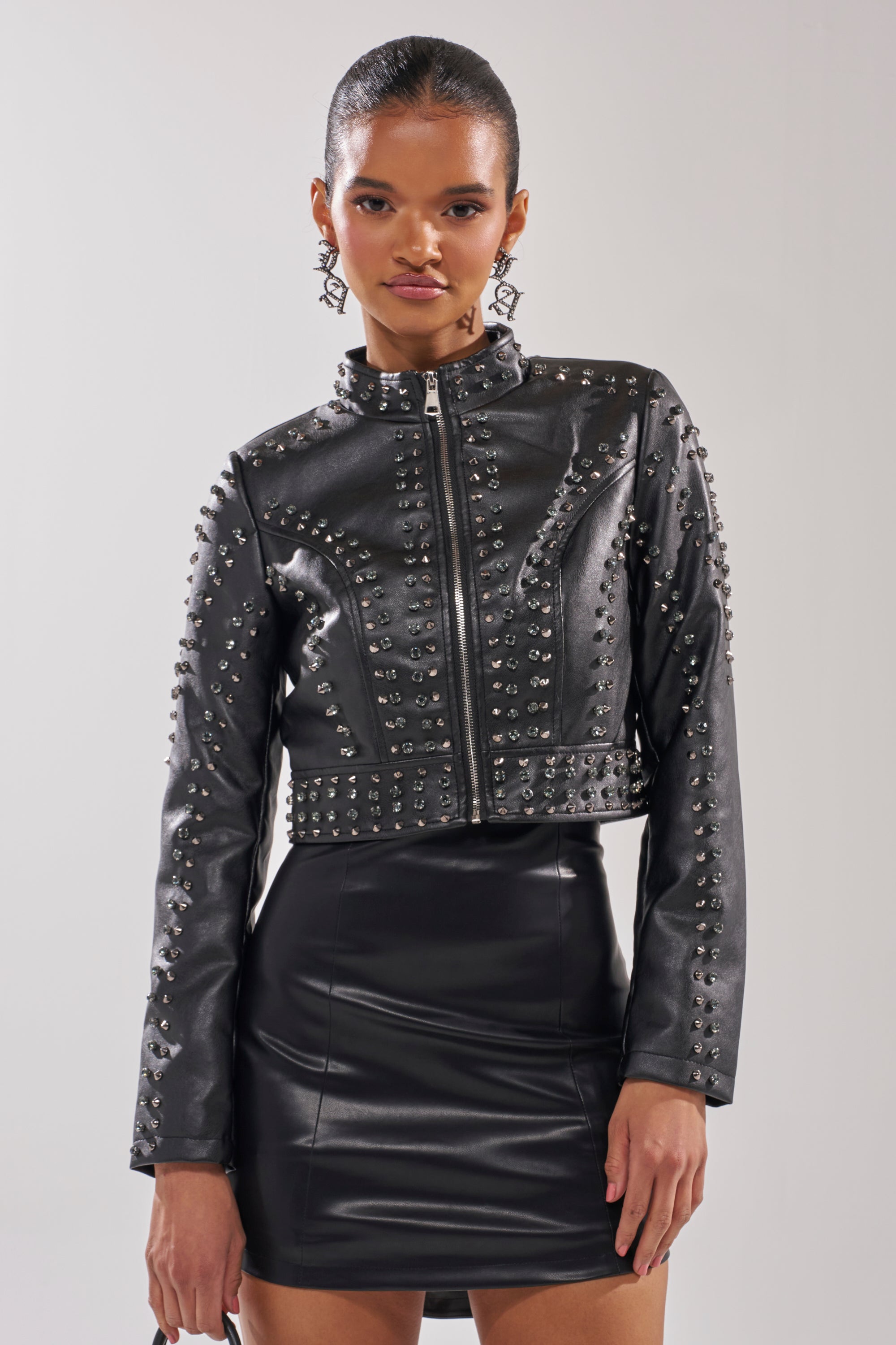 A woman in the SYLVIE STUDDED MOTO black studded moto jacket and a matching faux leather mini skirt stands against a plain light gray background, with slicked-back hair and statement earrings. 3