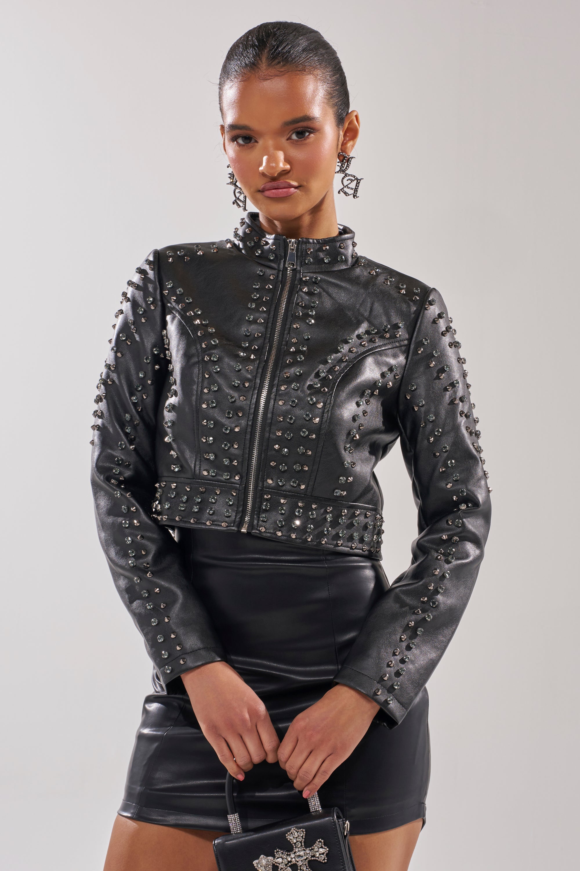 A woman wears the SYLVIE STUDDED MOTO jacket with a black leather mini skirt, holds a small black handbag, and completes her look with statement earrings and wet-look hair. 4