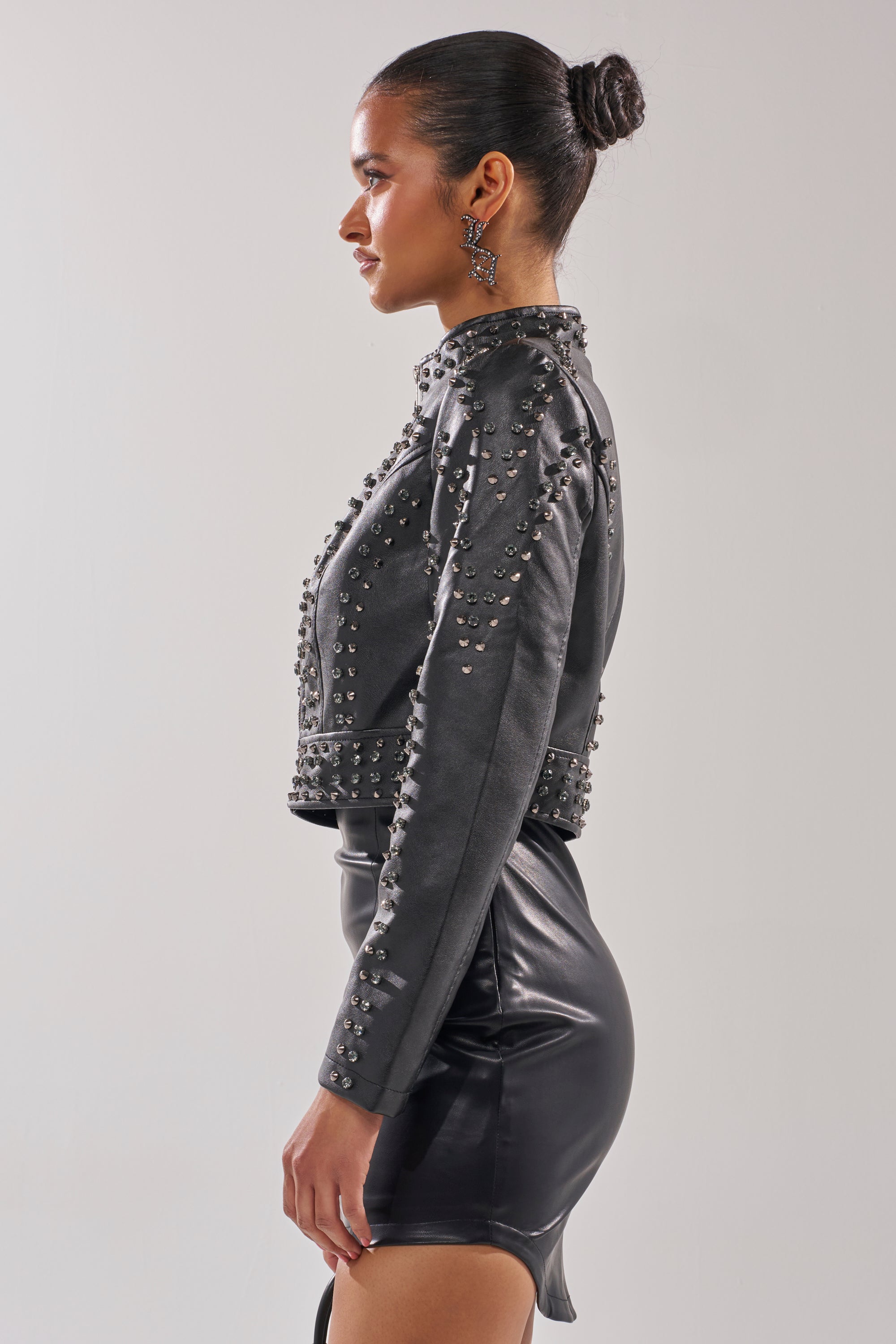 A woman in profile poses against a plain background, wearing the SYLVIE STUDDED MOTO jacket and a black leather mini skirt. Her hair is styled in a bun, and she completes the look with statement earrings. 5