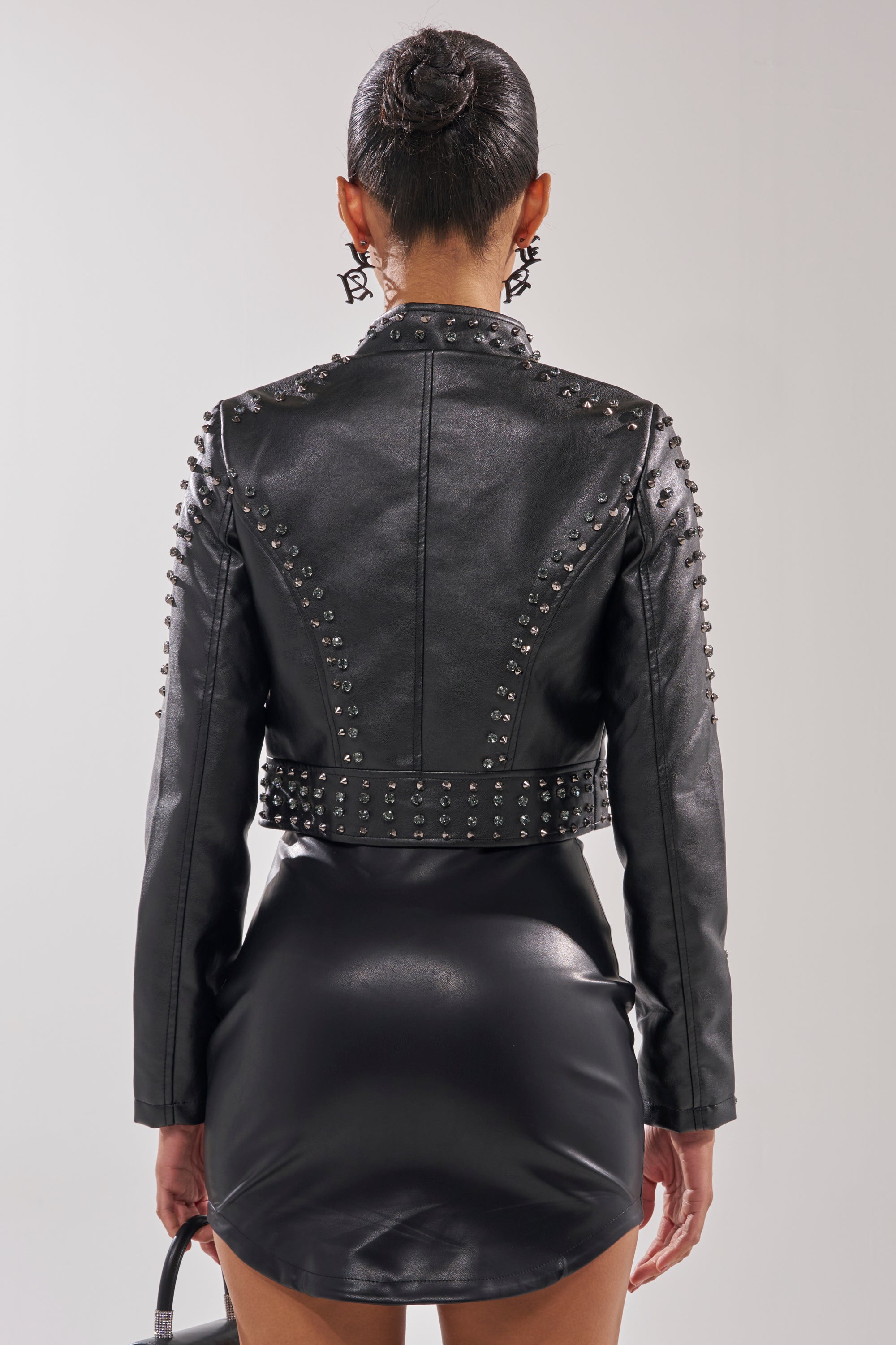 A person with hair in a sleek bun wears the SYLVIE STUDDED MOTO jacket and a matching short skirt, holding a small handbag and photographed from behind against a plain background. 6