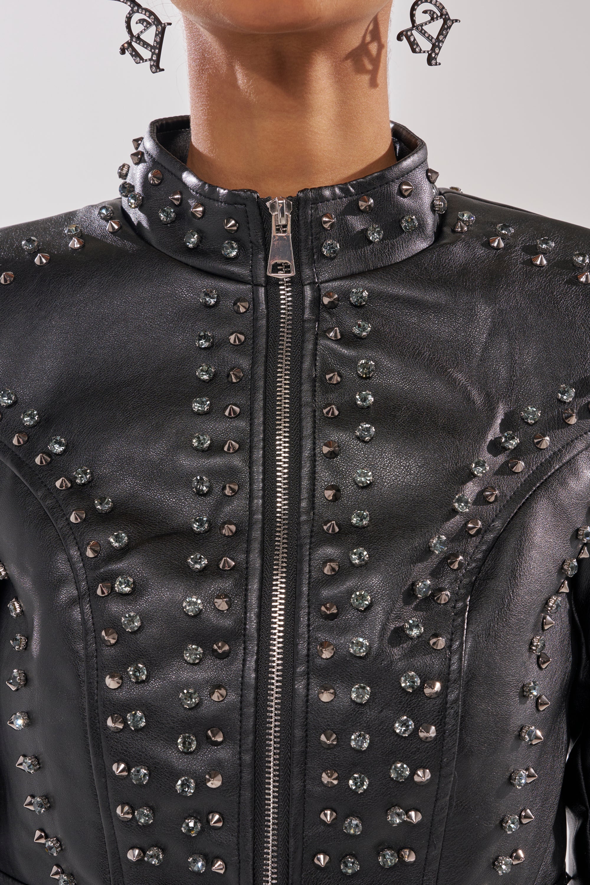 Close-up of a person wearing the SYLVIE STUDDED MOTO—a black moto jacket with front zipper, decorated with rows of metallic and gem-like studs. The shot shows the lower face, neck, and letter-shaped earrings. 8
