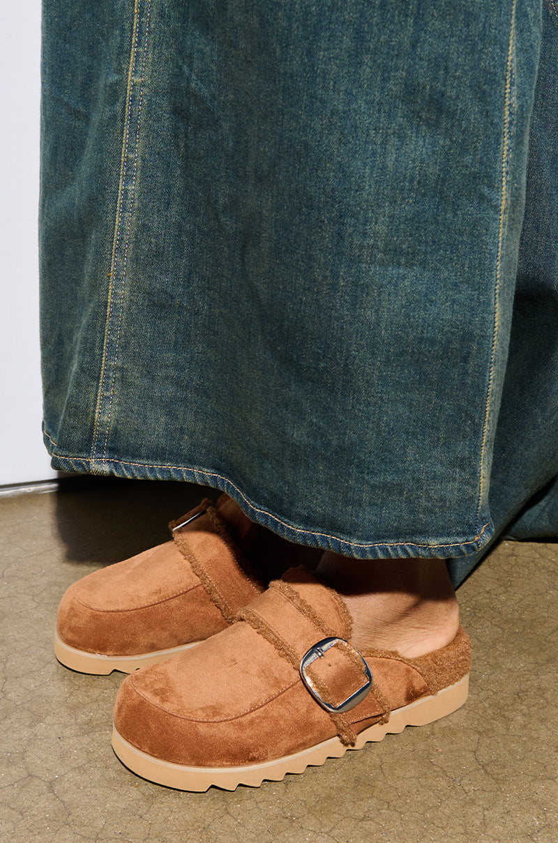 A person in blue denim jeans stands on a concrete floor, wearing AZALEA WANG SIMAH CHESTNUT SLIPPER CLOGS with a silver buckle and thick sole.