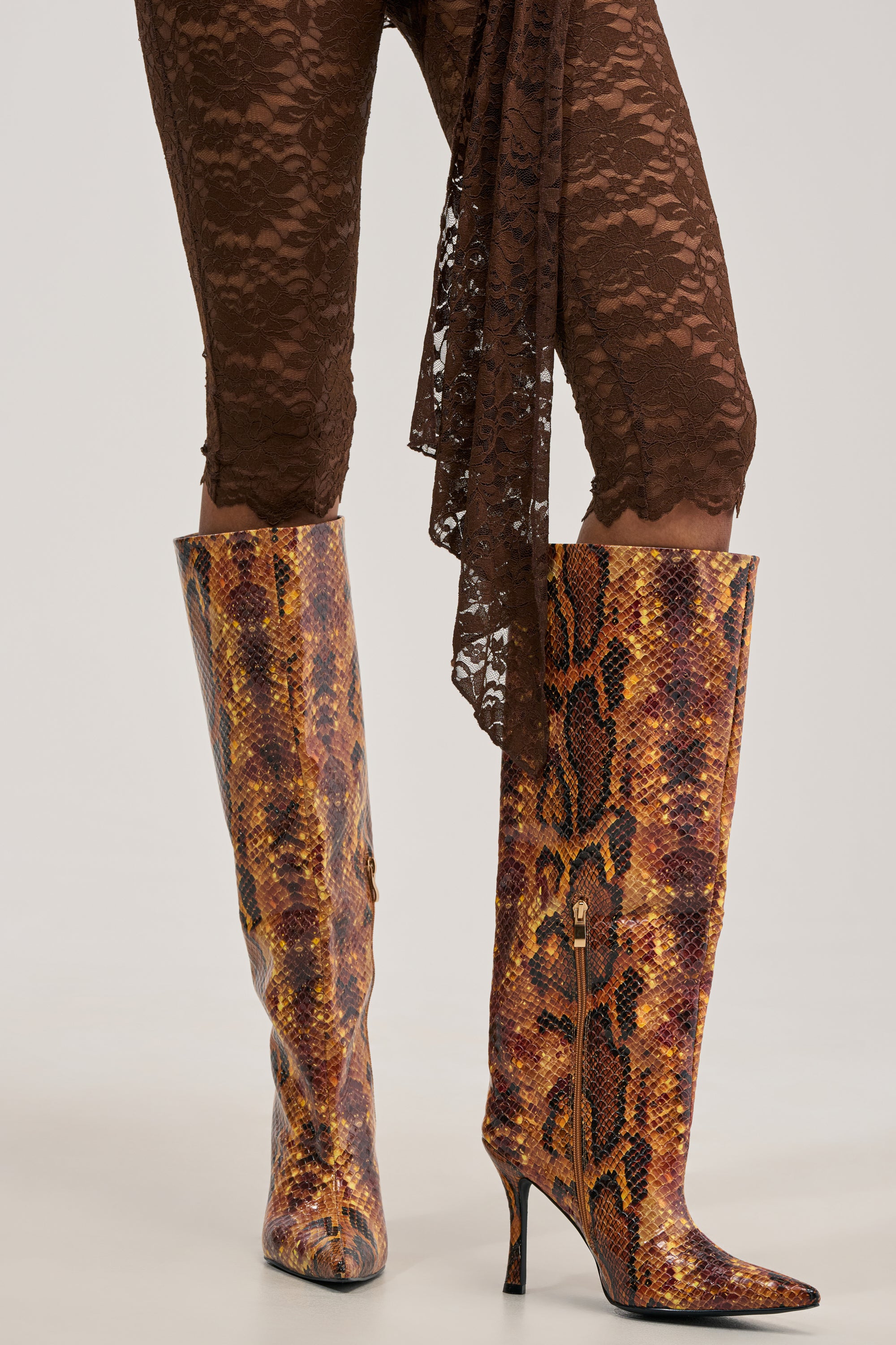 A person wearing brown lace pants and knee-high AZALEA WANG SIMONE ORANGE SNAKE STILETTO BOOTS featuring a bold brown and orange snakeskin pattern.
