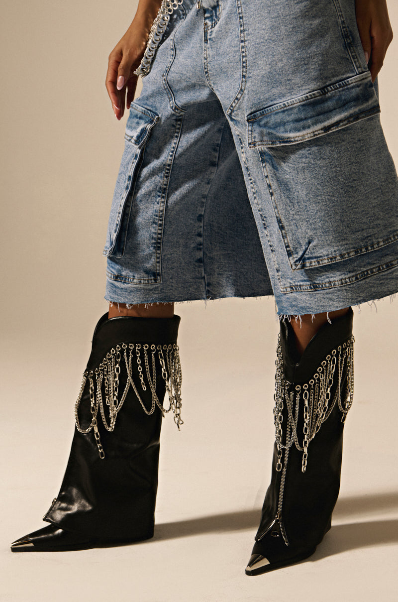 A close-up shows a person wearing knee-length distressed denim shorts with large pockets and fringed edges. They pair them with AZALEA WANG SINISTER BLACK WESTERN CHUNKY BOOTS, featuring metallic chains draping over the top, against a plain, light-colored background.