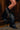 A close-up of a person wearing the AZALEA WANG SINISTER BLACK WESTERN CHUNKY BOOT, featuring metallic chains, standing indoors on a wooden floor against dark wood paneling.