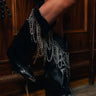 A close-up of a person wearing the AZALEA WANG SINISTER BLACK WESTERN CHUNKY BOOT, featuring metallic chains, standing indoors on a wooden floor against dark wood paneling.
