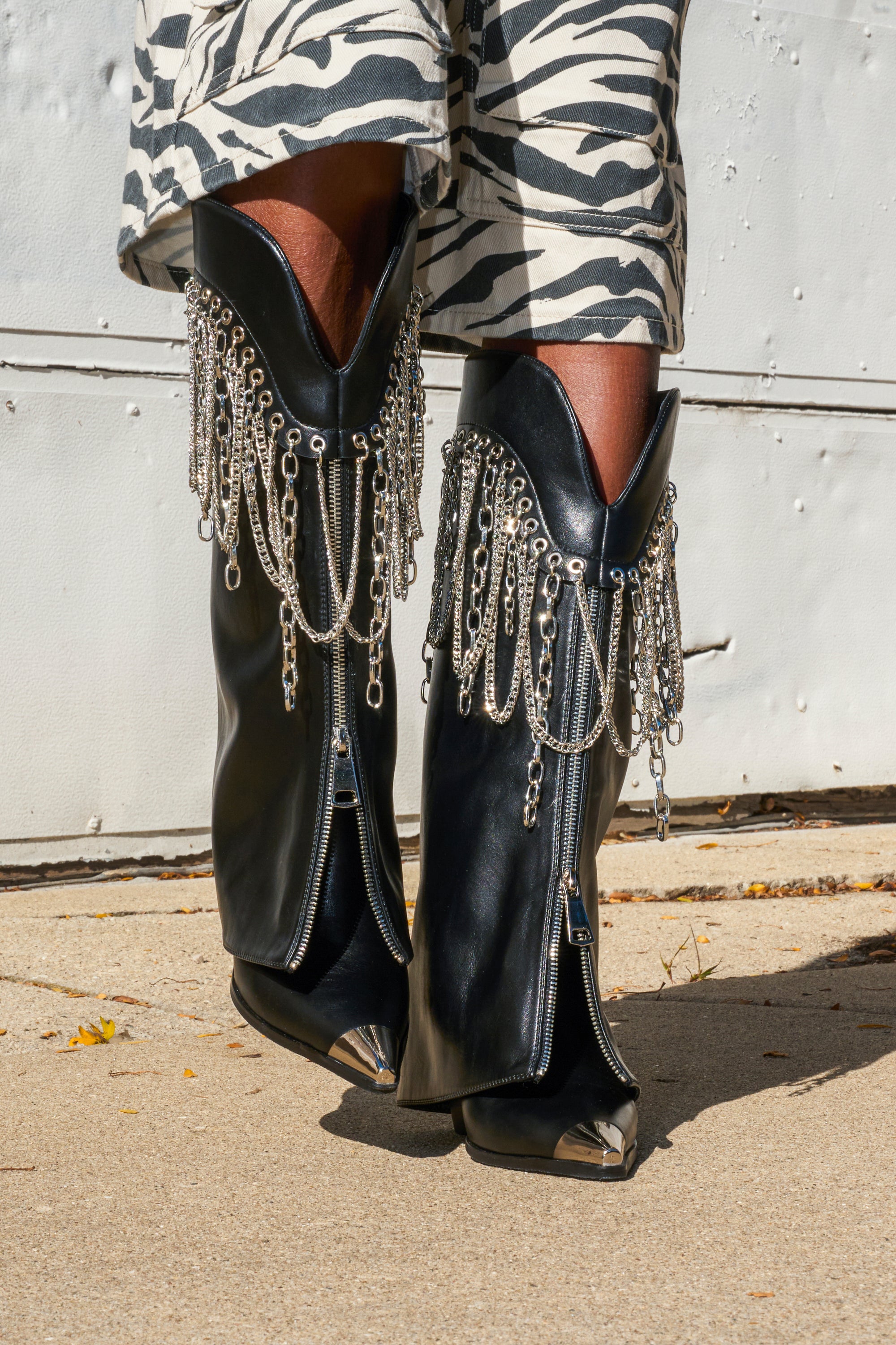 Close-up of a person wearing the AZALEA WANG SINISTER BLACK WESTERN CHUNKY BOOT with silver chain accents and metal toe caps, paired with zebra-print pants, standing on a concrete sidewalk.