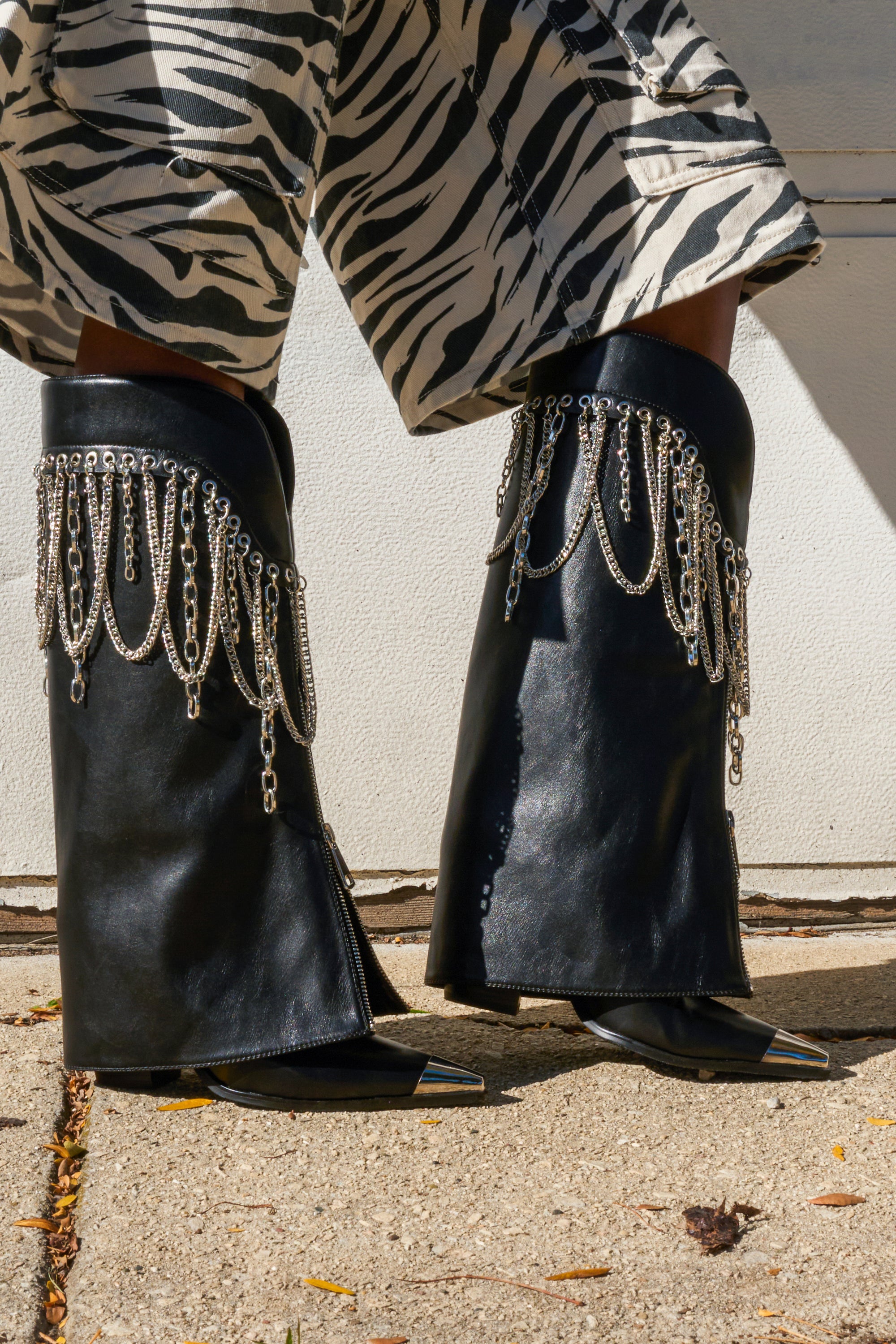 A person wearing wide-legged zebra-print pants and the AZALEA WANG SINISTER BLACK WESTERN CHUNKY BOOT with silver chain fringes, standing on a sunlit concrete sidewalk.