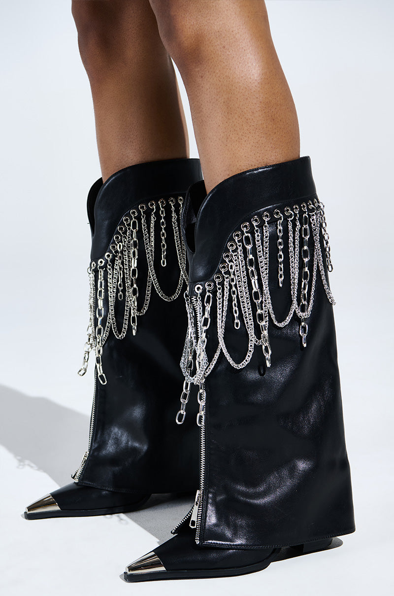 AZALEA WANG SINISTER BLACK WESTERN CHUNKY BOOT in black faux leather features silver chain fringe, metallic toe caps, decorative side zippers, and elegantly draped metallic chains on the top. Posed on a light background.